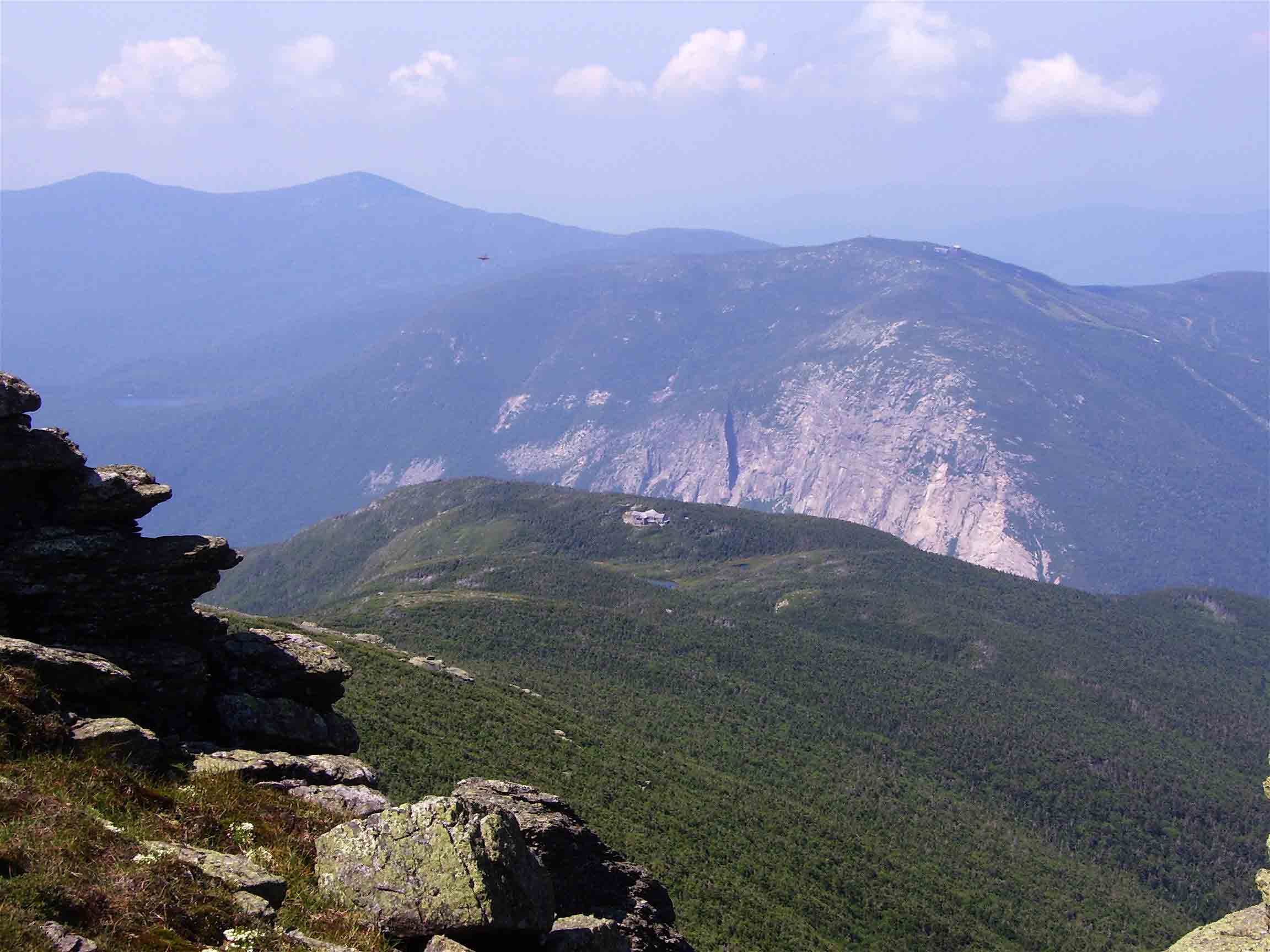 mm 21.3 - View to the west from the ridge of Mt. Lafayette. Greenleaf Hut (not on AT) is in center of picture. The mountain with the spectacular cliff is Cannon Mt. on the other side of Franconia Notch. The two large peaks on the left in the distance are North and South Kinsman, both of which the AT crosses.  Courtesy dlcul@conncoll.edu